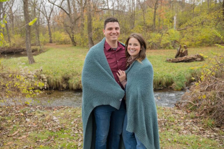 Couple standing outdoors by a creek, wrapped together in a soft teal Saranoni blanket. The lush fall scenery and cozy blanket highlight the comfort and warmth of the moment, showcasing a favorite gift idea for relaxation and connection.