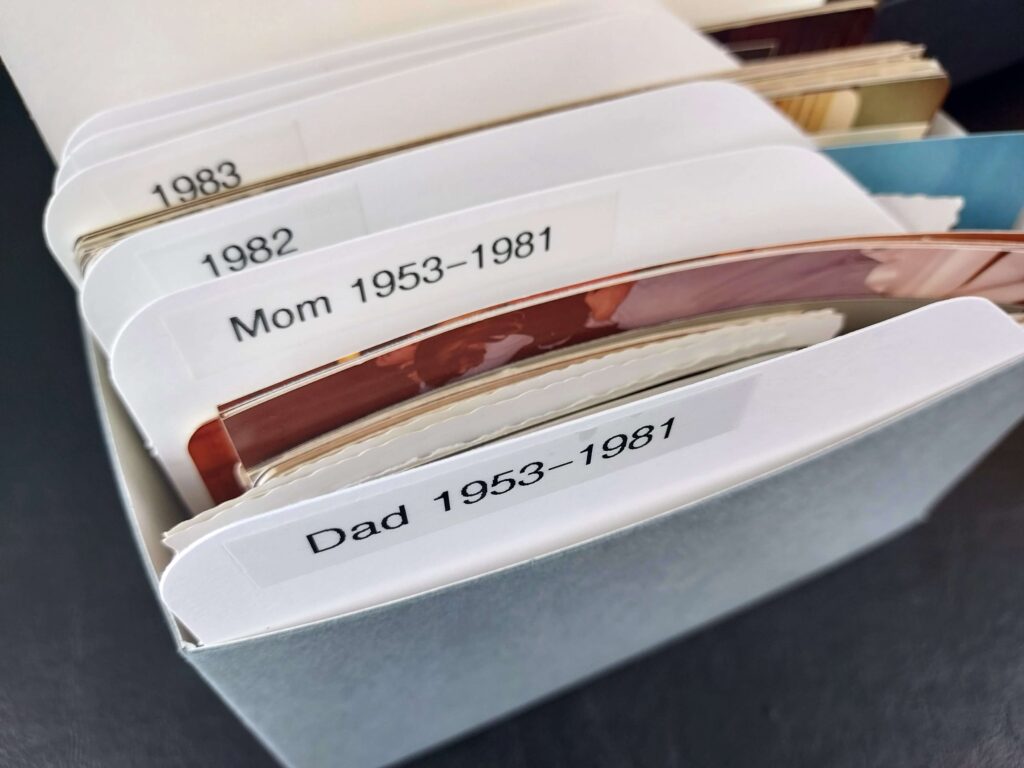 Close-up of an organized photo storage box with labeled dividers separating printed photographs by year and family members, including labels such as '1983,' '1982,' 'Mom 1953-1981,' and 'Dad 1953-1981.' A practical example of a system for preserving and categorizing family memories.