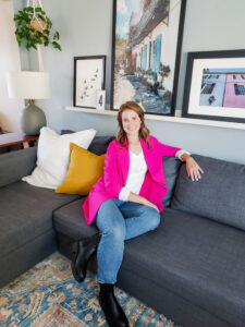 Professional home organizer in Ann Arbor, MI, sitting on a gray sectional couch wearing a bright pink jacket, with wall art and modern decor in the background.