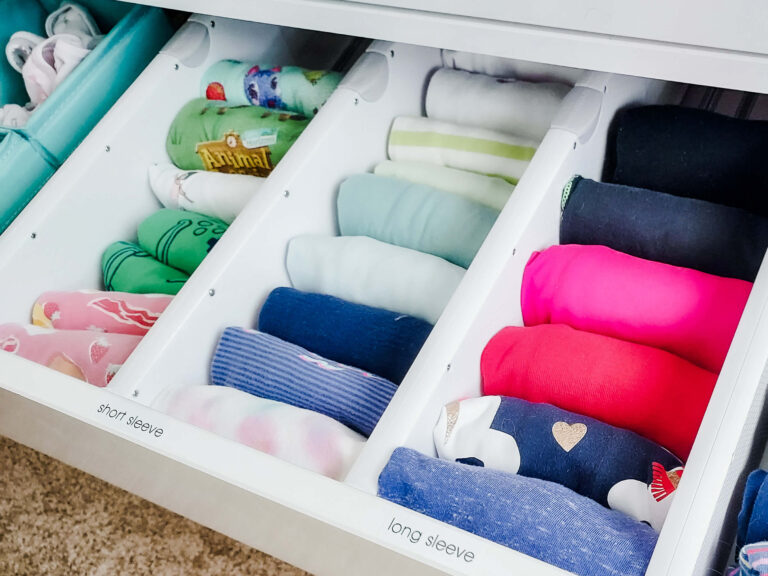 A neatly organized drawer featuring children's long sleeve shirts, divided into sections by adjustable drawer dividers. The shirts are file folded and arranged by color for easy access and visibility, demonstrating Ashley's expertise in maximizing space and creating functional, organized storage solutions.